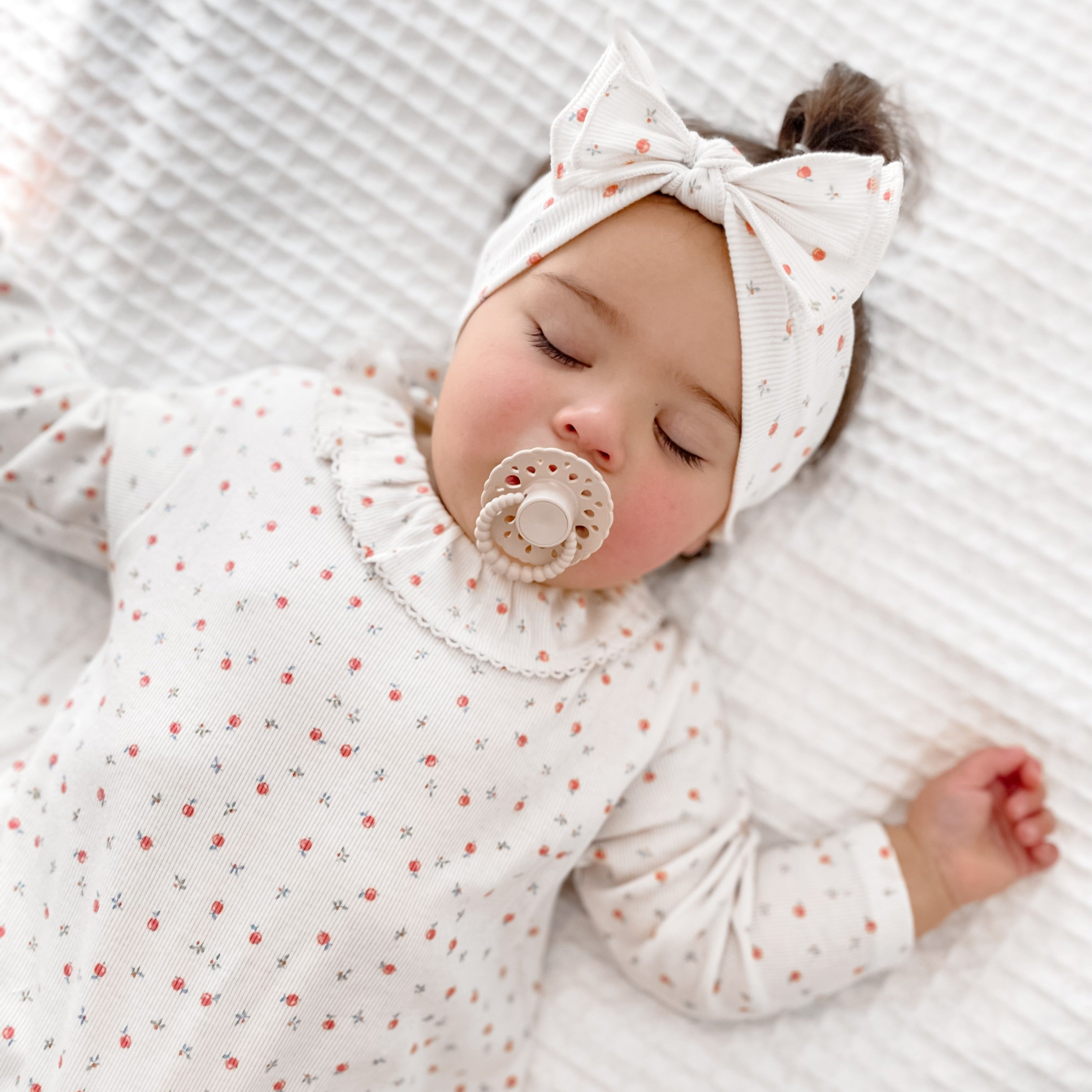 Sleeping baby girl wearing white floral outfit and headband using a oatmeal scalloped edge bloom cmc dummy with bubble handle