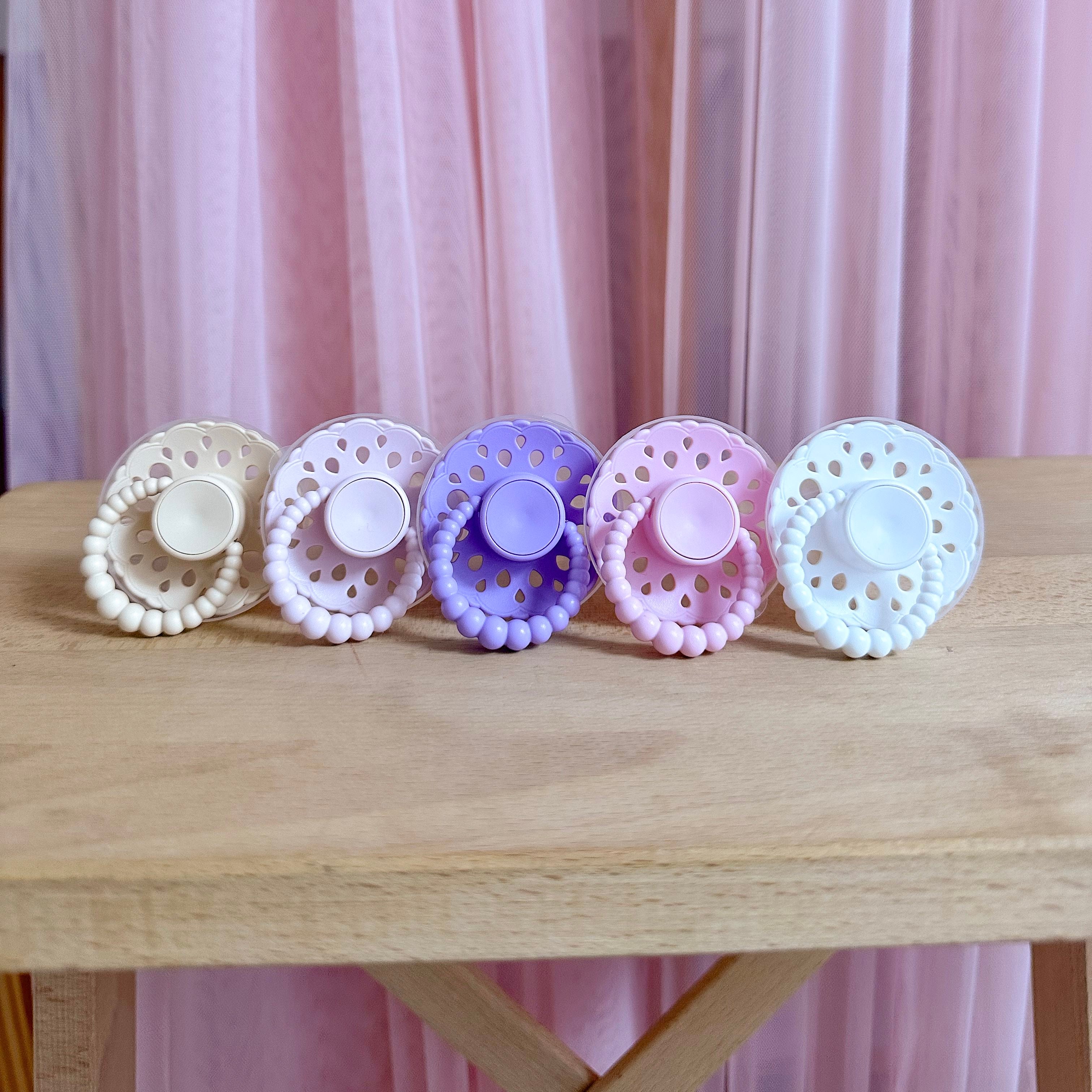Set of bloom baby dummies in pastel colours including oatmeal, blush, lavender, pink and white displayed on a wooden table