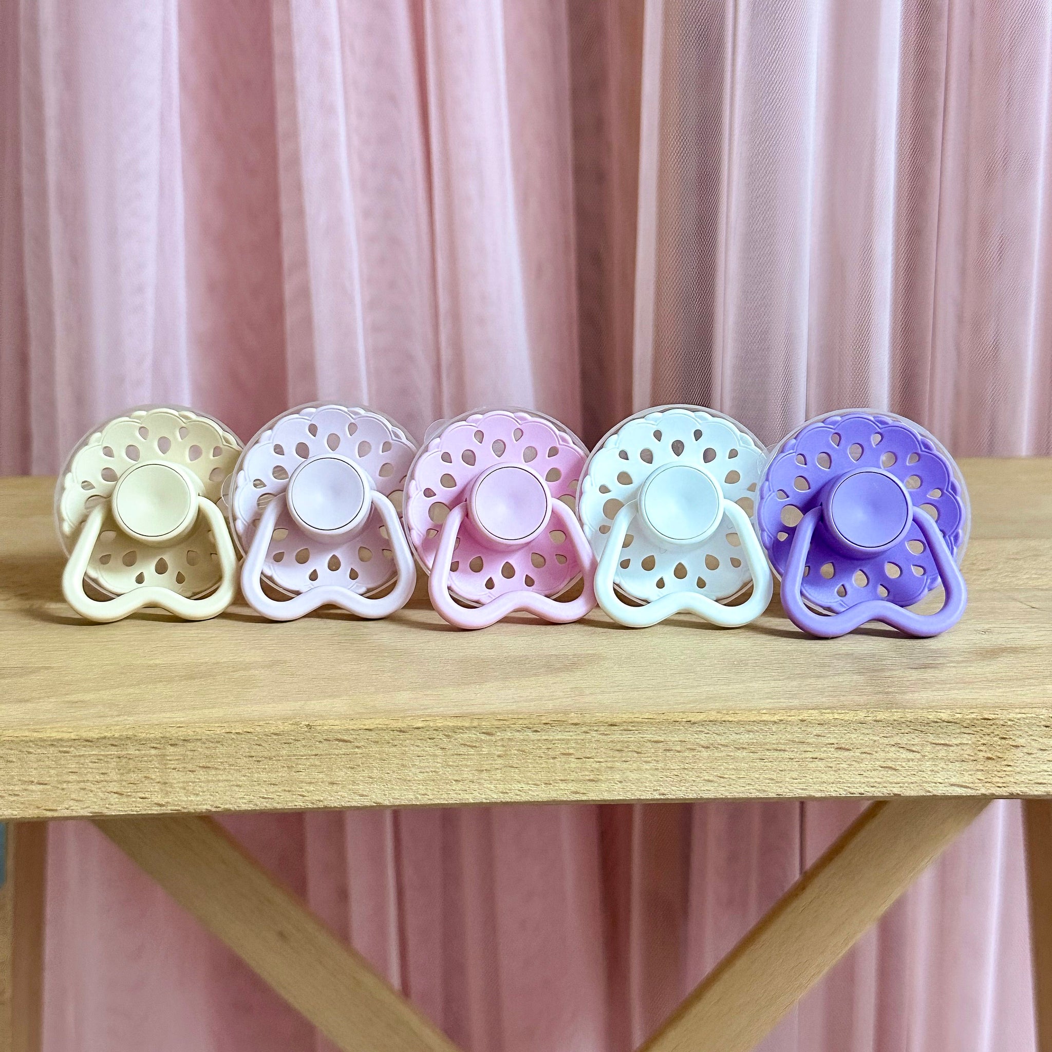 Set of bloom baby dummies in pastel colours including oatmeal, blush, lavender, pink and white with hold me handles displayed on a wooden table