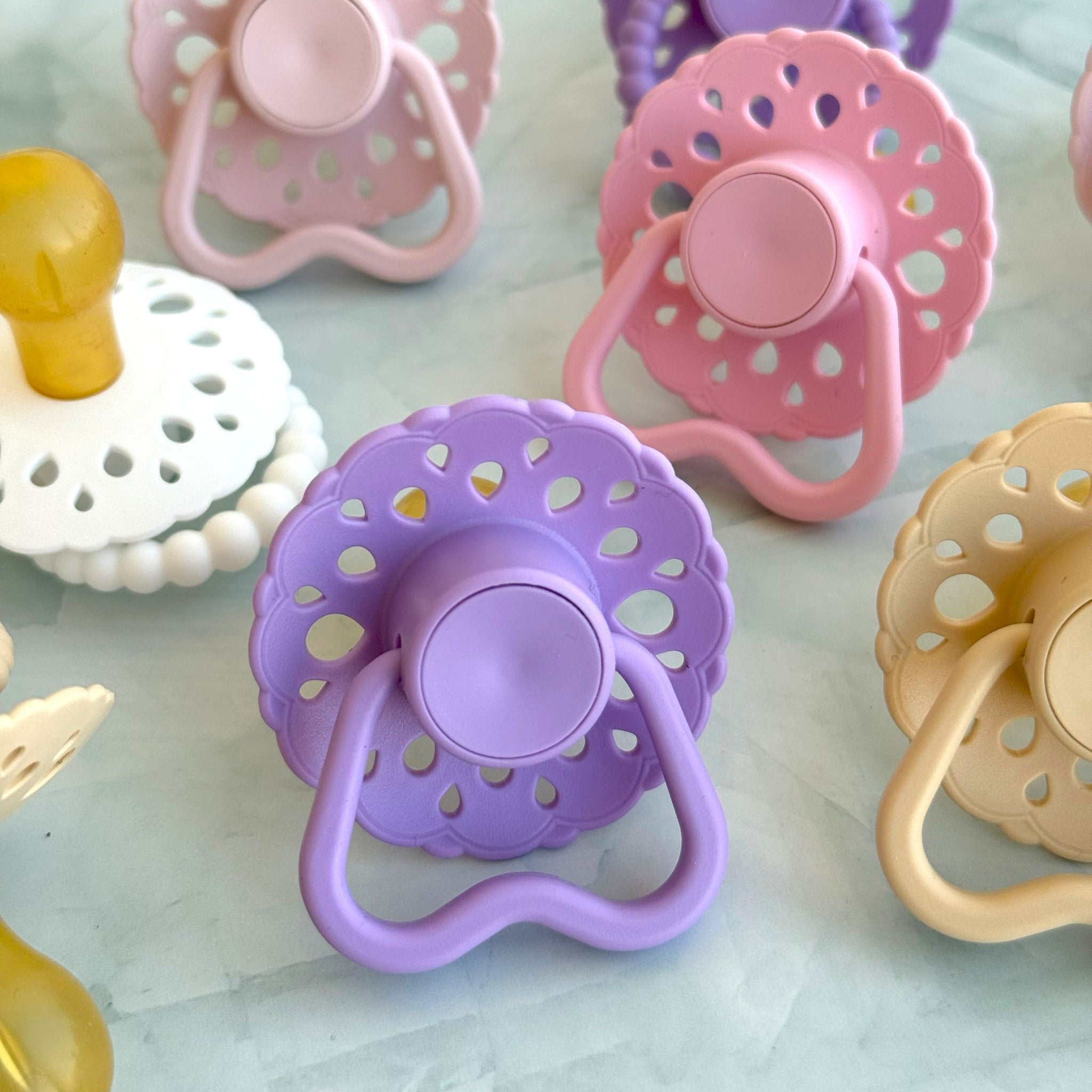 Set of bloom baby dummies in pastel colours including oatmeal, blush, lavender, pink and white with hold me handles displayed on a wooden table