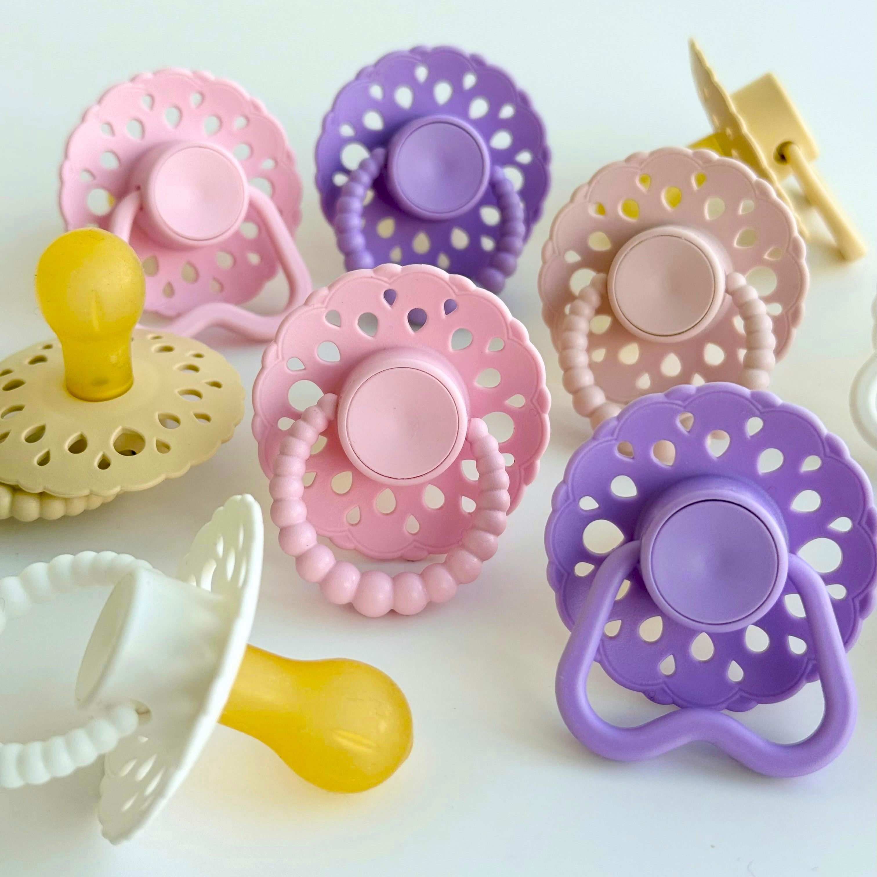 Set of bloom baby dummies in pastel colours including oatmeal, blush, lavender, pink and white featuring hold me and bubble handles displayed on a wooden table