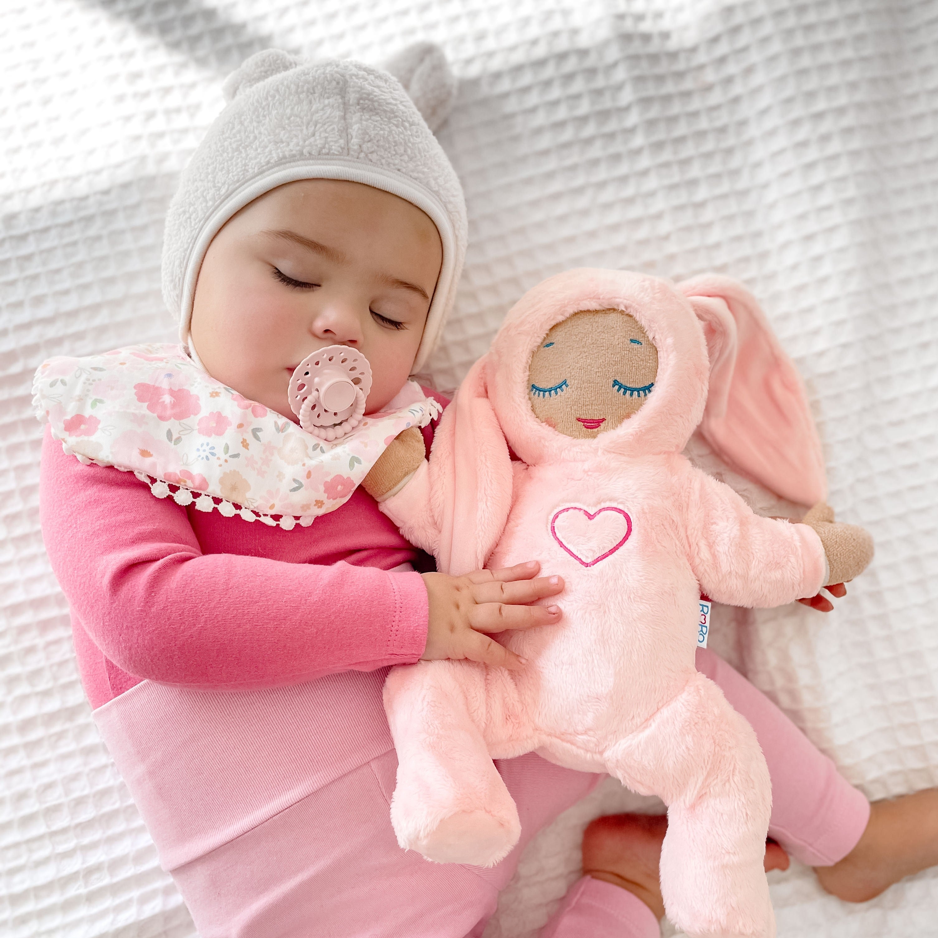 Sleeping baby girl wearing pink outfit and beanie using a blush bloom cmc scalloped edge dummy with bubble handle while holding a soft plush toy.