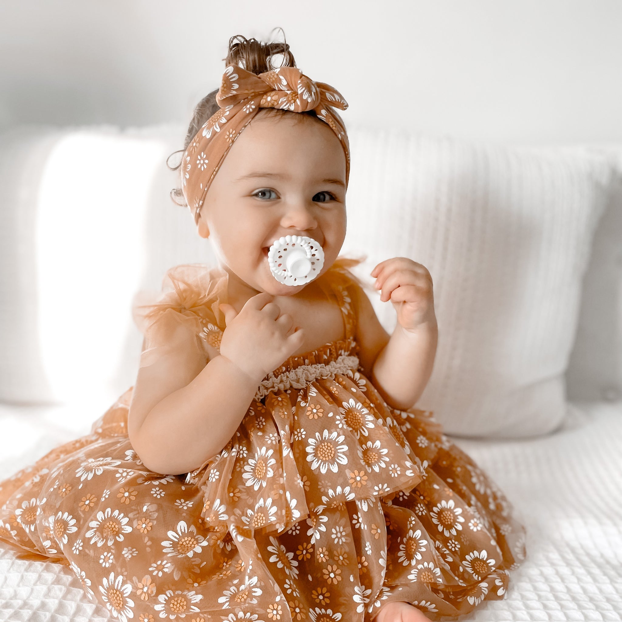 Smiling baby girl wearing a floral outfit and headband using a white bloom dummy with scalloped edge and bubble handle while sitting on a couch
