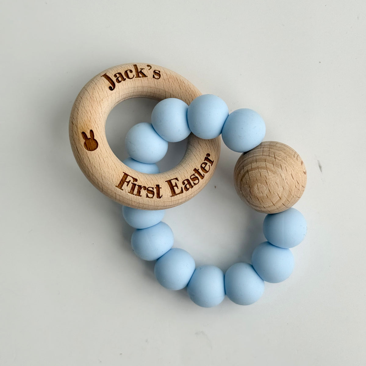 Personalised wooden and blue silicone baby teether engraved with “Jack’s First Easter” featuring a natural wood ring and soft silicone beads