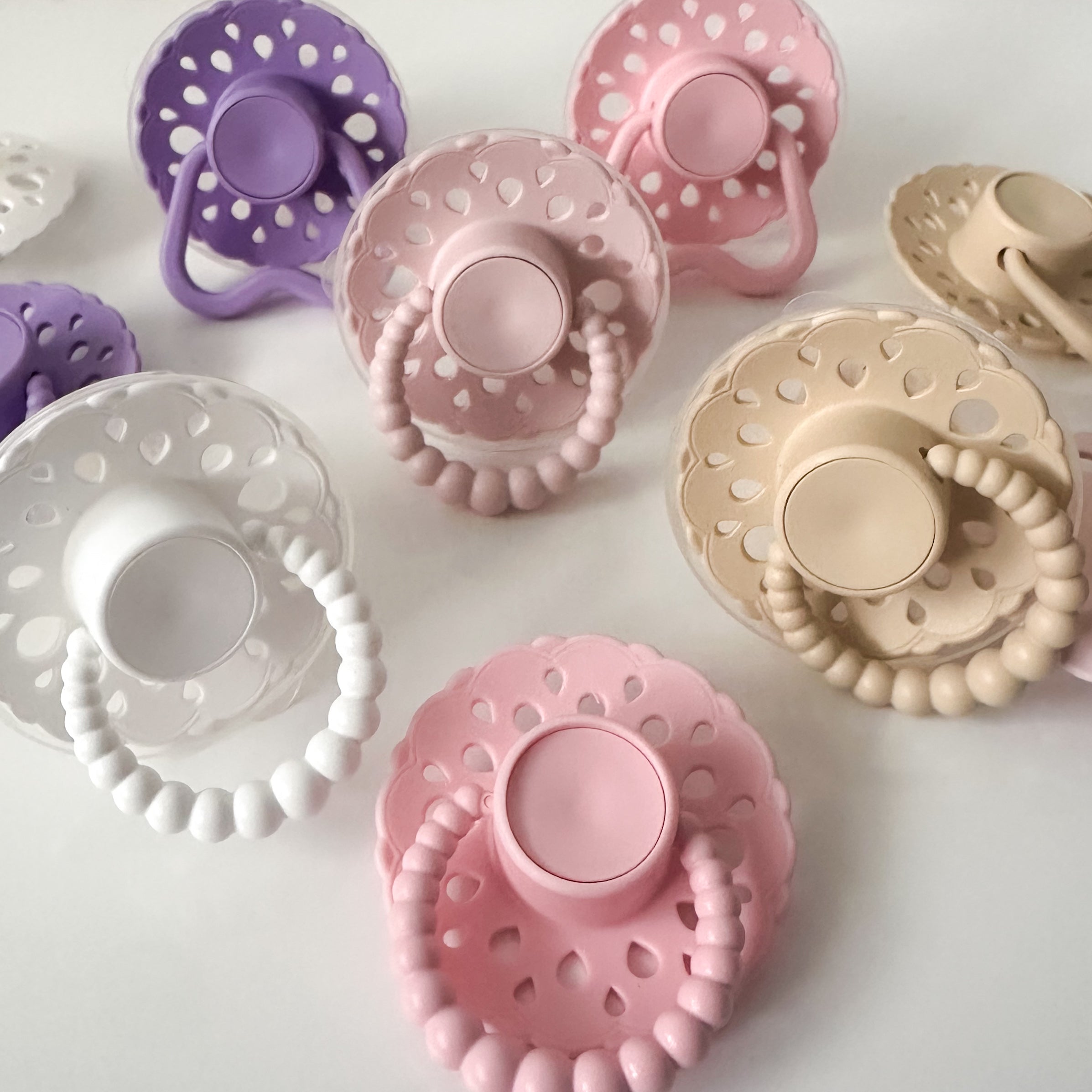 Set of bloom baby dummies in pastel colours including oatmeal, blush, lavender, pink and white displayed on a wooden table