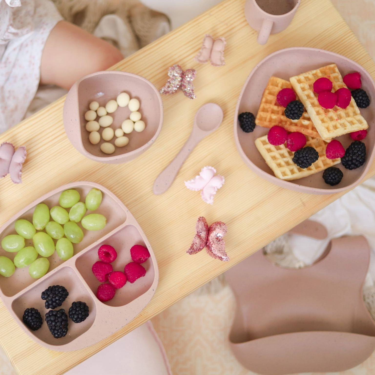 Silicone baby feeding set with divided bento plate, regular plate, bowl, spoon, cup and waffles with fruit on a wooden table