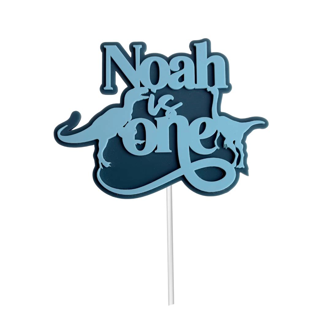 Personalised first birthday cake topper with the name Noah is One, featuring a dinosaur design in bluestone and candy floss acrylic