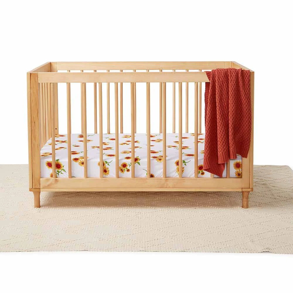 Baby cot with sunflower print fitted sheet and rust knitted blanket in a wooden nursery crib